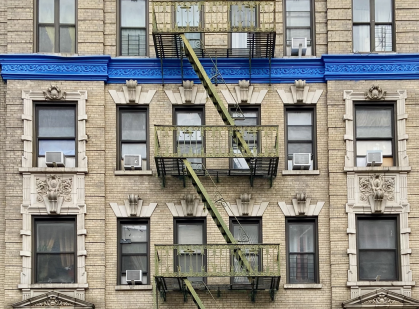 The facade of a residential prewar brick building with a green fire escape and a bright blue cornice in West Harlem, Manhattan, New York City